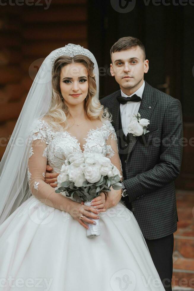 hermoso novio y encantador novia juntos cerca moderno de madera casas en el parque. retrato. magnífico vestir con un largo tren. elegante joven novio foto