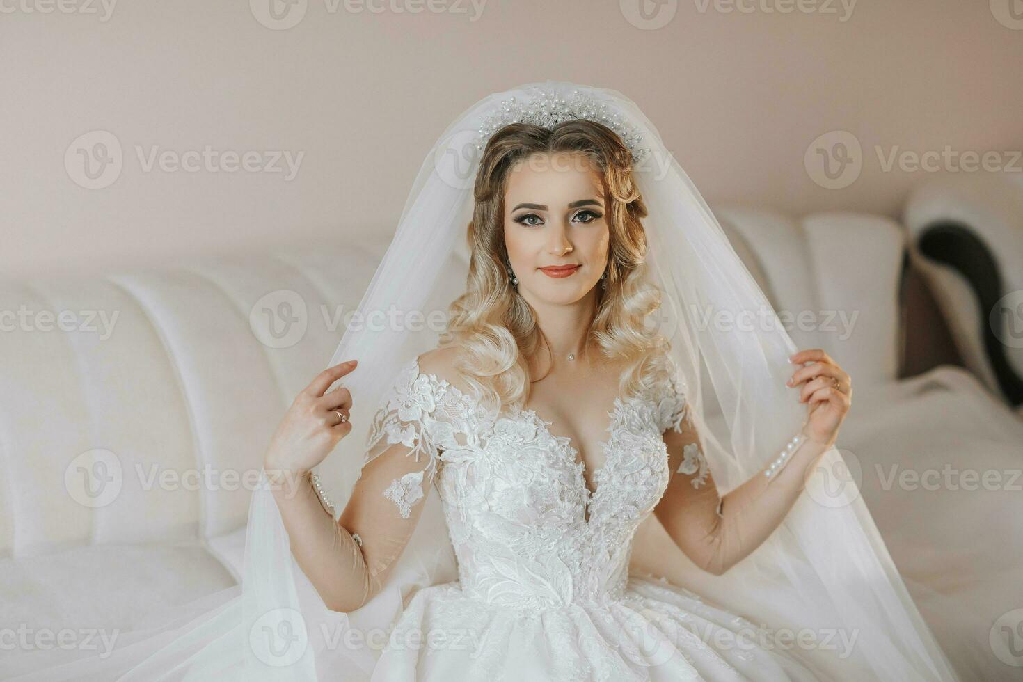 the bride in a white wedding dress. happy beautiful young woman in white traditional wedding dress in her room on leather sofa holding veil photo