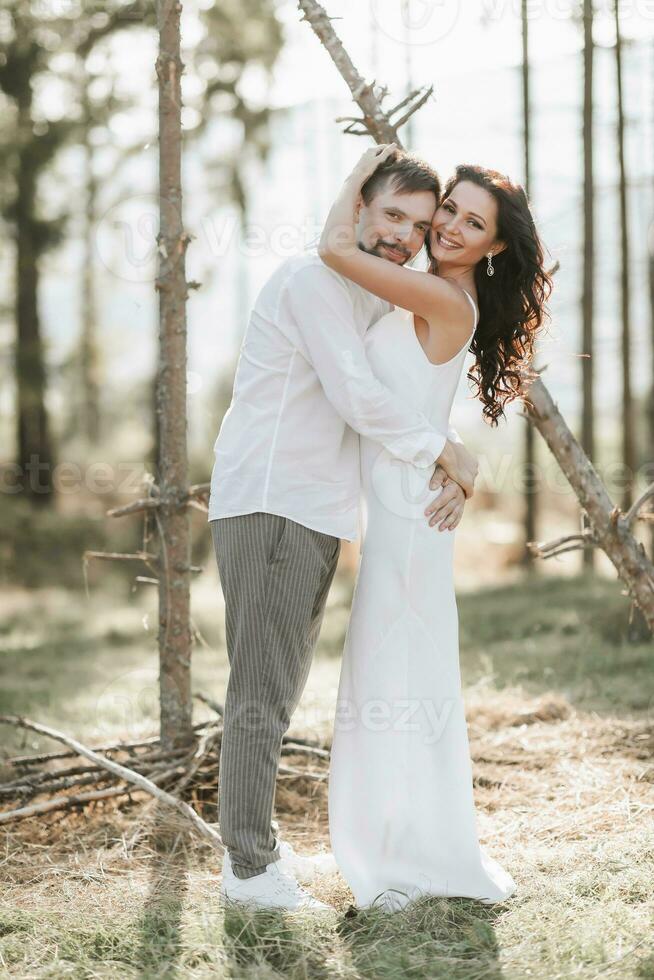 elegante novio en blanco camisa y linda morena novia en blanco vestir en bosque cerca Boda de madera arco. Boda retrato de recién casados. foto