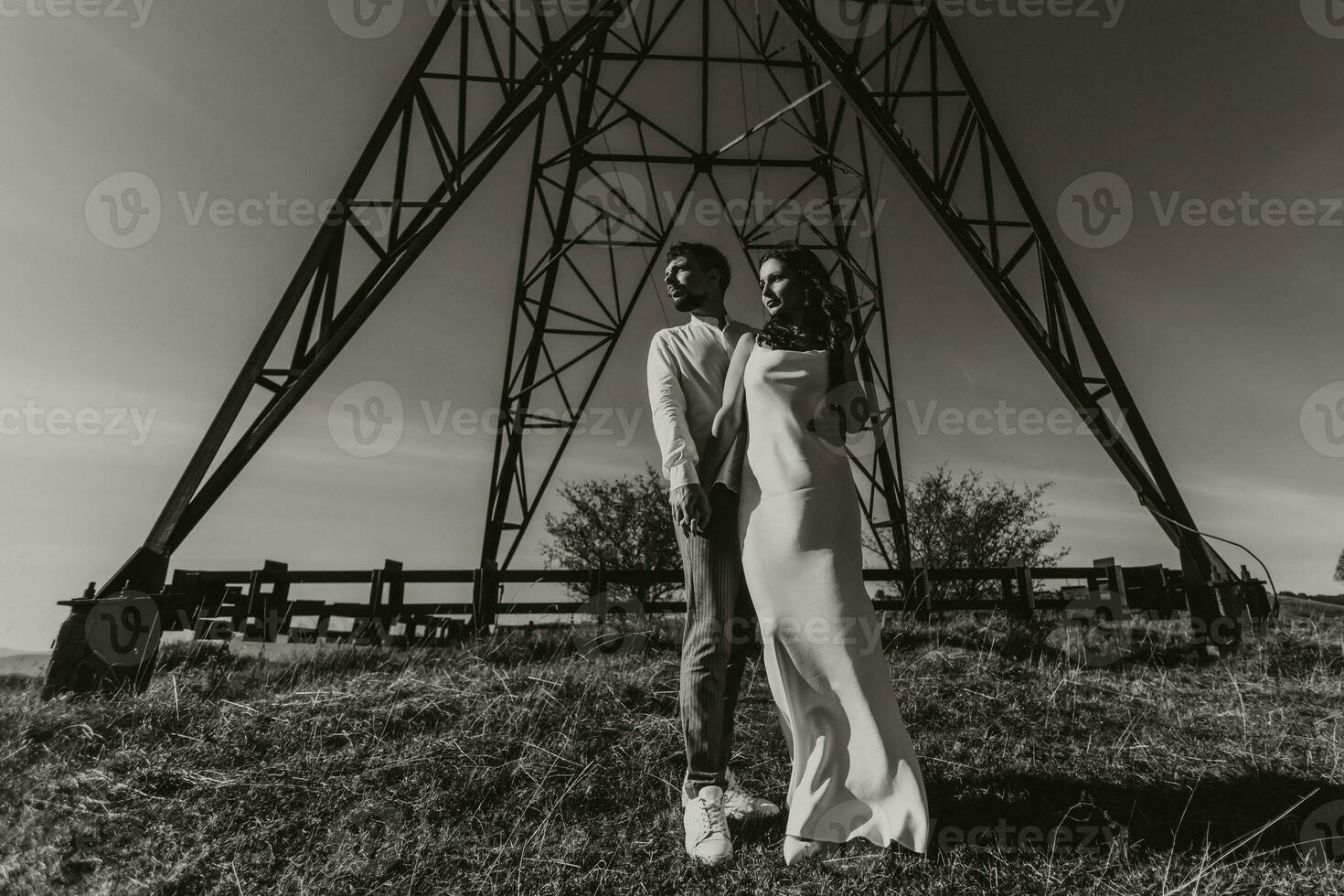 Stylish model couple in the mountains in summer. A young boy and girl in a white silk dress are standing near large structures of power lines. Black and white contrast photo