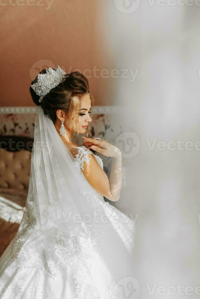 The bride in a beautiful dress by the window. Photo from the back of the Wedding. wedding bouquet Morning of the bride. An artistic wedding. A happy bride is waiting for the groom.