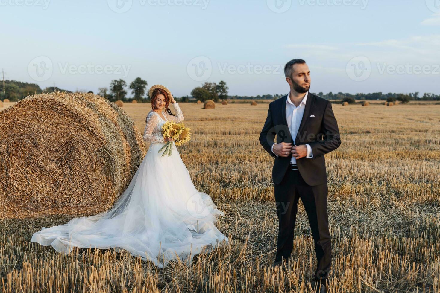 Wedding portrait of the bride and groom. The groom is standing in front of the bride, adjusting his jacket. Red-haired bride in a long dress with a bouquet of sunflowers. Stylish groom. Summer photo