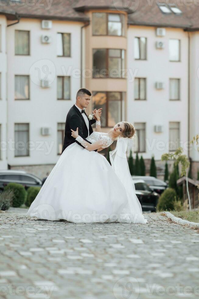 Wedding portrait. A groom in a black suit and a blonde bride dance a waltz against the background of buildings. Long dress in the air. Photo session in nature. Beautiful hair and makeup