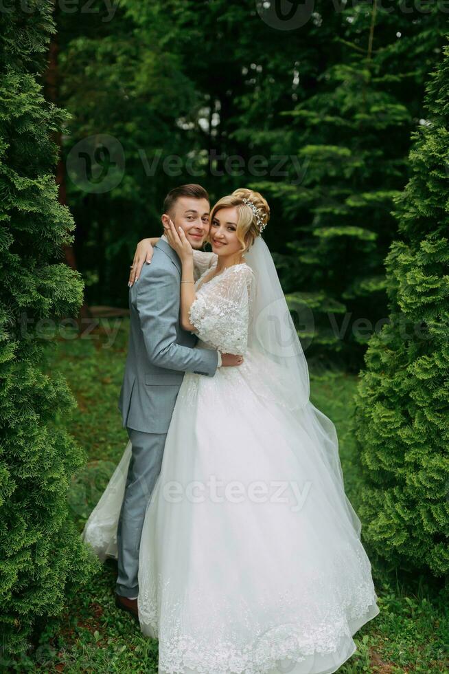 Portrait of the bride and groom standing on the background of green trees, embracing. The bride smiles sincerely. Stylish groom. Fashion and style. Beautiful bride photo