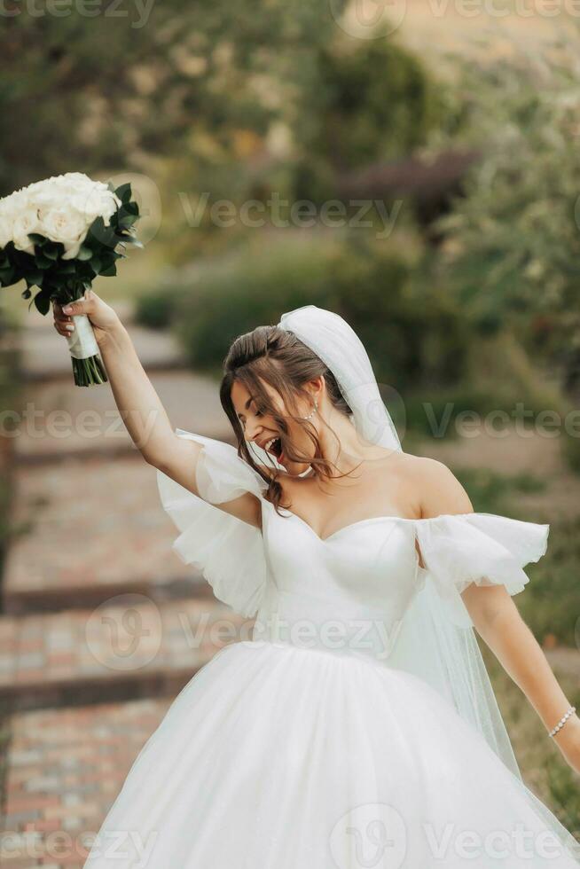 Portrait of the bride in nature. A brunette bride in a white long dress, holding a bouquet of white roses, poses, looking into the lens, smiling sincerely. on the background of the tree. Curly hair. photo