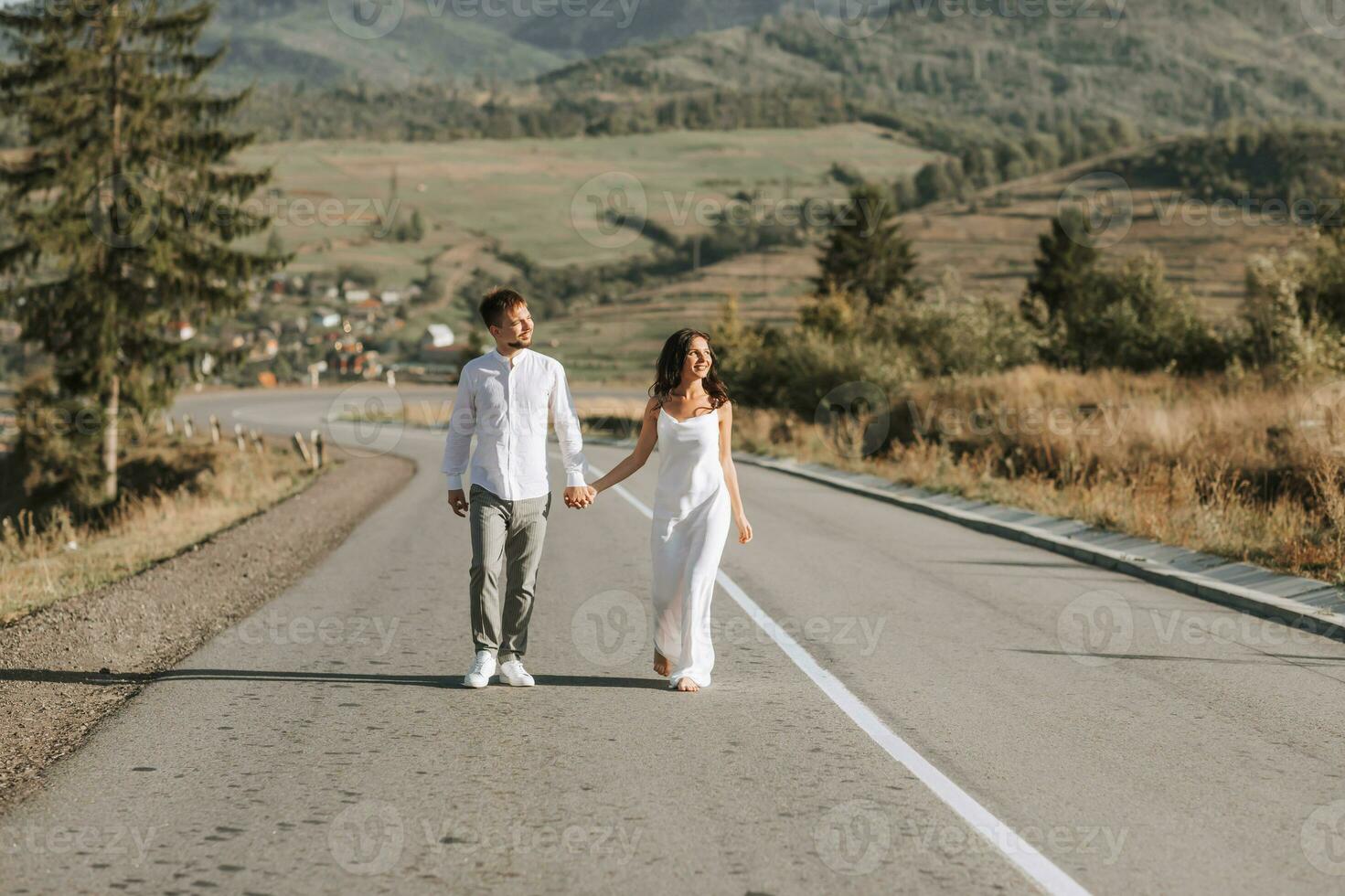 A stylish groom in a white shirt and a cute brunette bride in a white dress are walking on an asphalt road against the background of a forest and mountains. Wedding portrait of newlyweds. photo