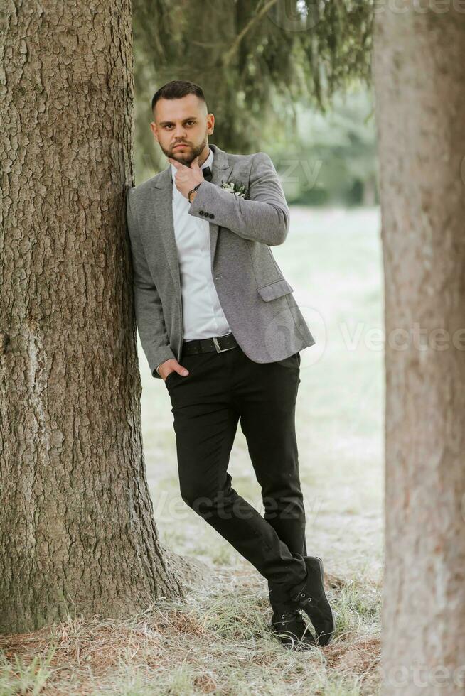 elegante acicalar, propensión su hombro en contra un árbol, enfatiza su barba, mira dentro el cámara lente. retrato foto