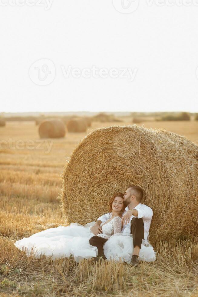 Wedding portrait. The bride and groom are sitting in an embrace on the ground, near a bale of hay. Red-haired bride in a long dress. Stylish groom. Summer. A sincere smile. On the background of hay photo