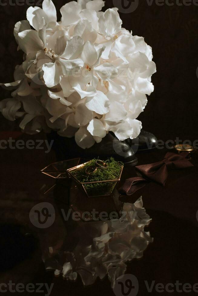 Morning of the groom and details, details of the groom on a dark background. men's watch, wedding rings in a glass box and wooden moss, a men's butterfly and a bouquet of white flowers photo