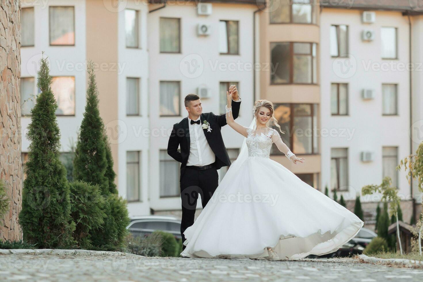 Wedding portrait. A groom in a black suit and a blonde bride dance a waltz against the background of buildings. Long dress in the air. Photo session in nature. Beautiful hair and makeup