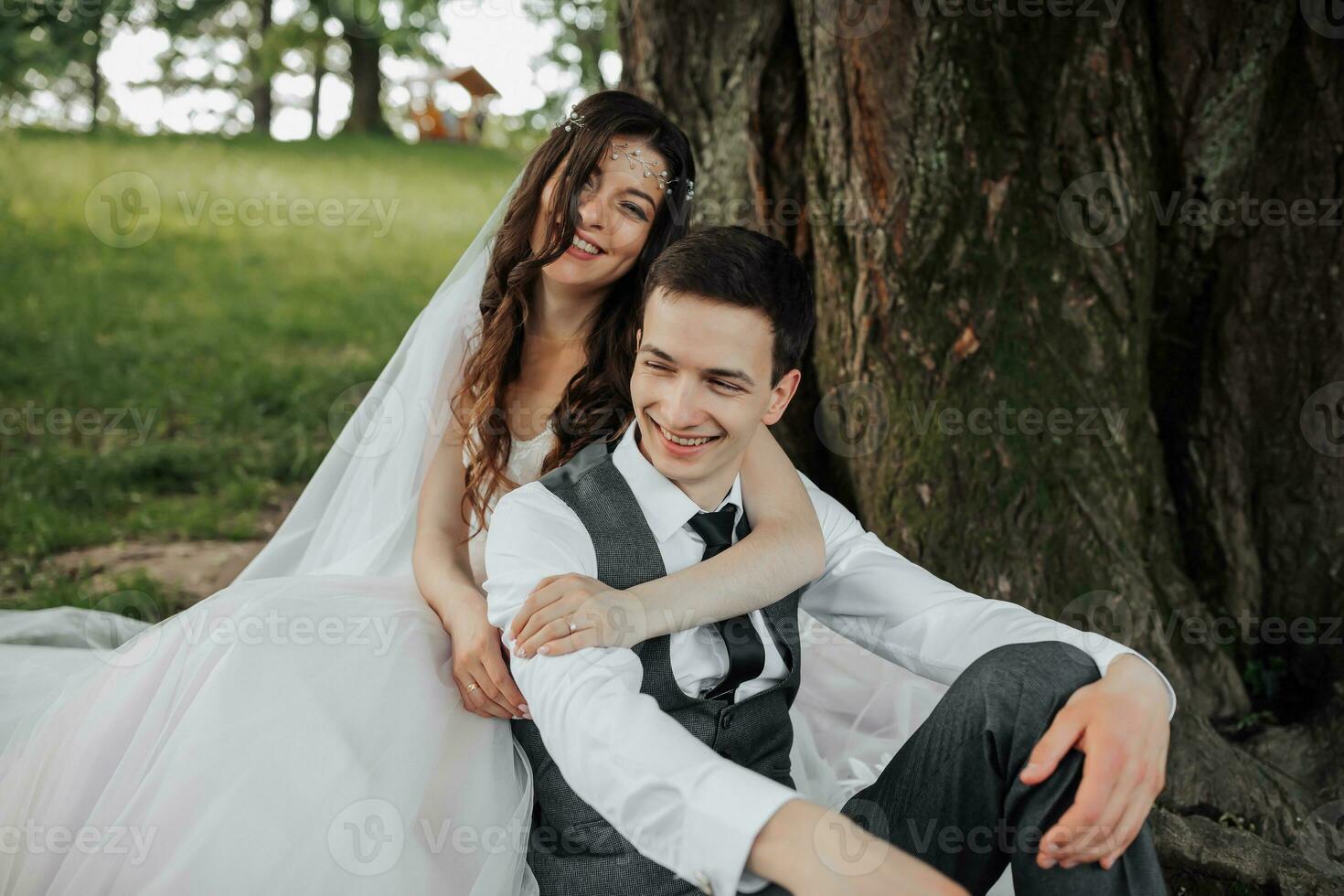 A beautiful bride with long curly hair in a chic dress hugs the groom, smiles, looking into the lens under a big tree. Portrait of the bride and groom. Spring wedding. Natural makeup photo