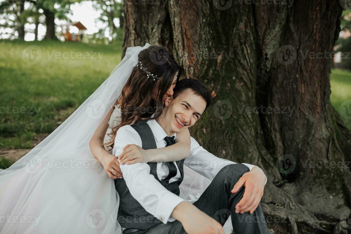 A beautiful bride with long curly hair in a chic dress hugs the groom, smiles, looking into the lens under a big tree. Portrait of the bride and groom. Spring wedding. Natural makeup photo
