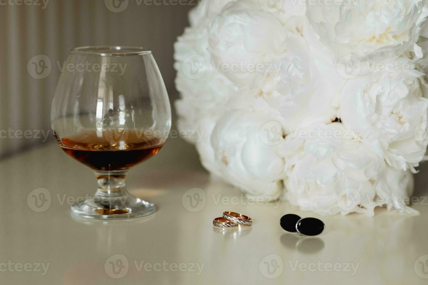 Accessories for the groom's wedding. A goblet with a drink, a white bouquet for the bride, gold rings, cufflinks on a gray table. Men's fashion photo