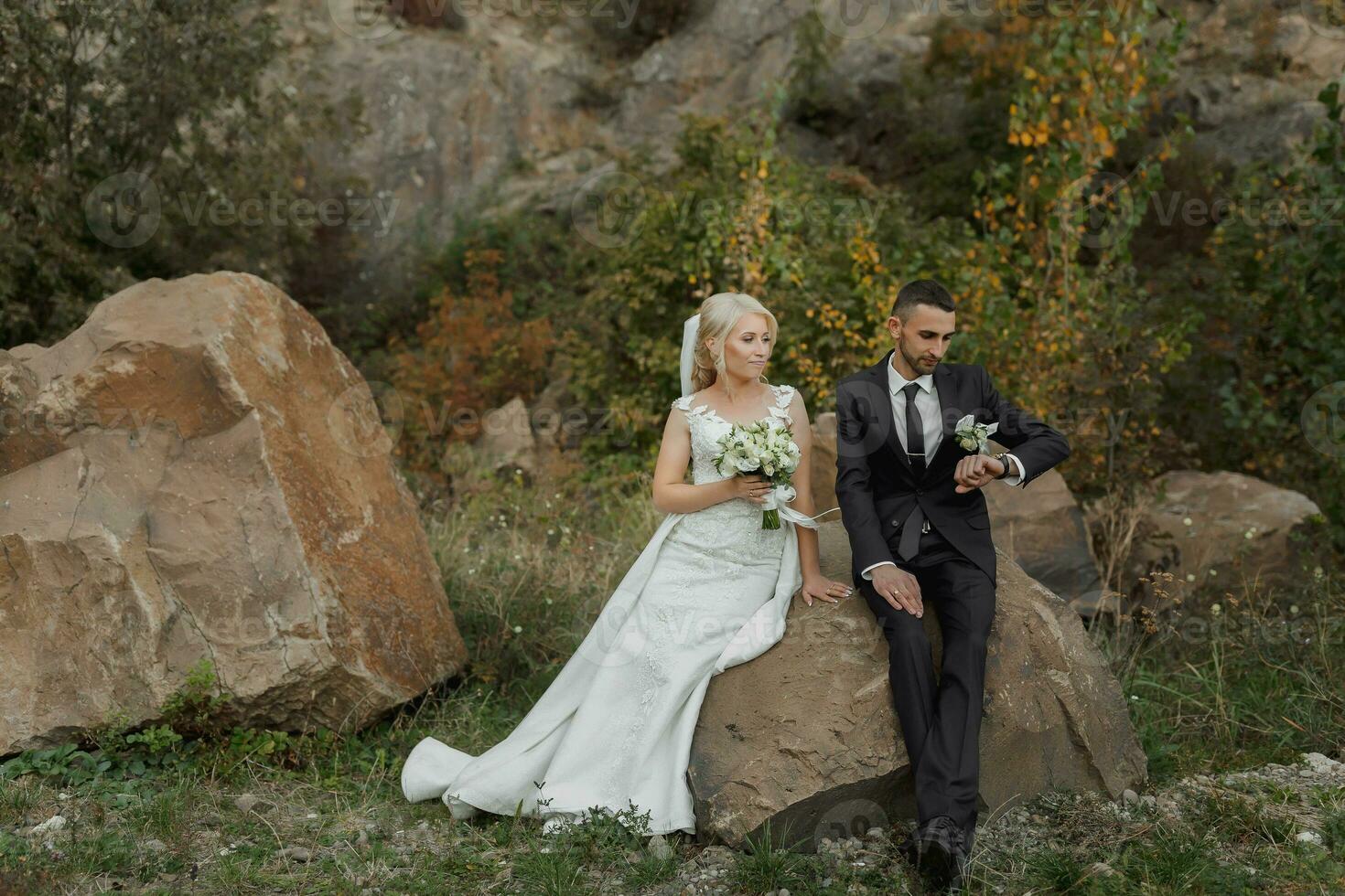 The bride and groom are standing on a rock high in the mountains next to a large stone. The bride is under a veil, the groom is hugging her shoulders photo