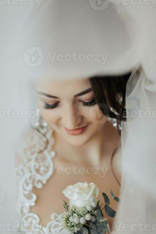 A brunette bride in a white dress holds her wedding bouquet, poses. French manicure. Open shoulders. Beautiful hands. Long veil. Morning of the bride. Details photo