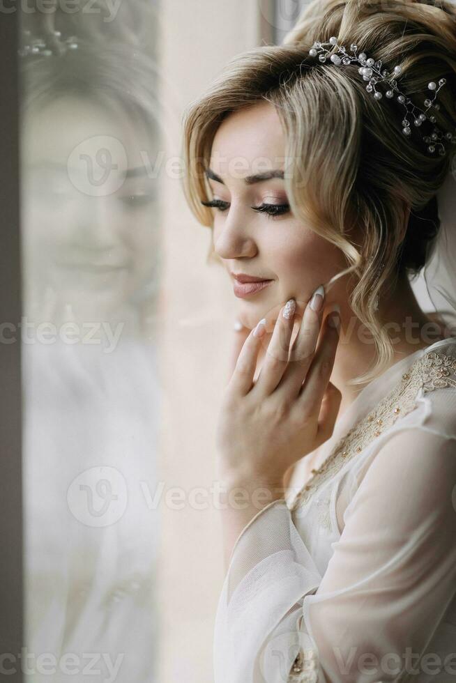 Blonde bride with elegant hairdo and tiara, wearing a robe, posing, looking at the camera, touching her face. Beautiful makeup. Elegant hairstyle. French manicure photo