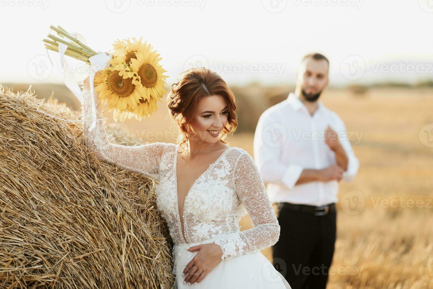 Wedding portrait of the bride and groom. The groom, tearing his shirt, stands behind the bride, near a bale of hay. Red-haired bride in a long dress with a bouquet of sunflowers. Stylish groom. Summer photo