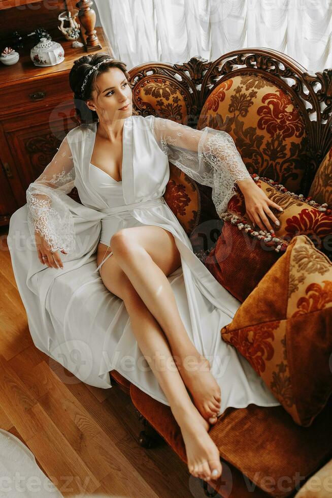 beautiful bride on a retro sofa in a robe with open legs, full length photo. Wedding hairstyle, light makeup photo