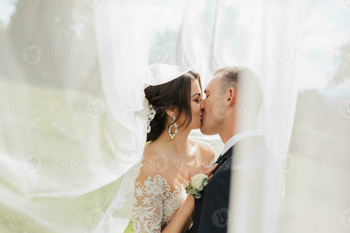 A curly-haired brunette bride in a white dress and a veiled groom embrace and kiss. Portrait of the bride and groom. Beautiful makeup and hair. Wedding in nature photo