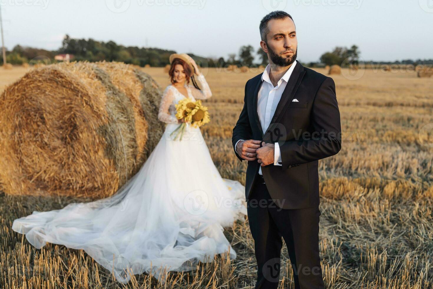 Wedding portrait of the bride and groom. The groom is standing in front of the bride, adjusting his jacket. Red-haired bride in a long dress with a bouquet of sunflowers. Stylish groom. Summer photo
