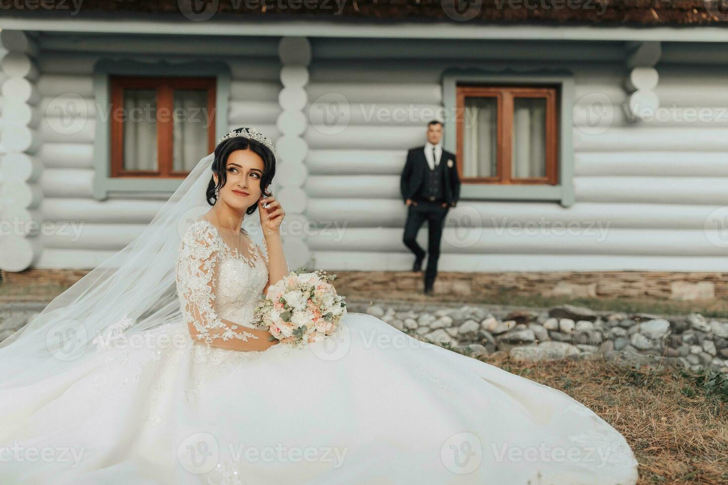 A brunette bride with a crown on her head in a white voluminous dress is posing, holding a bouquet in her hands, the groom is standing behind her leaning on a white hut. Wedding shooting photo