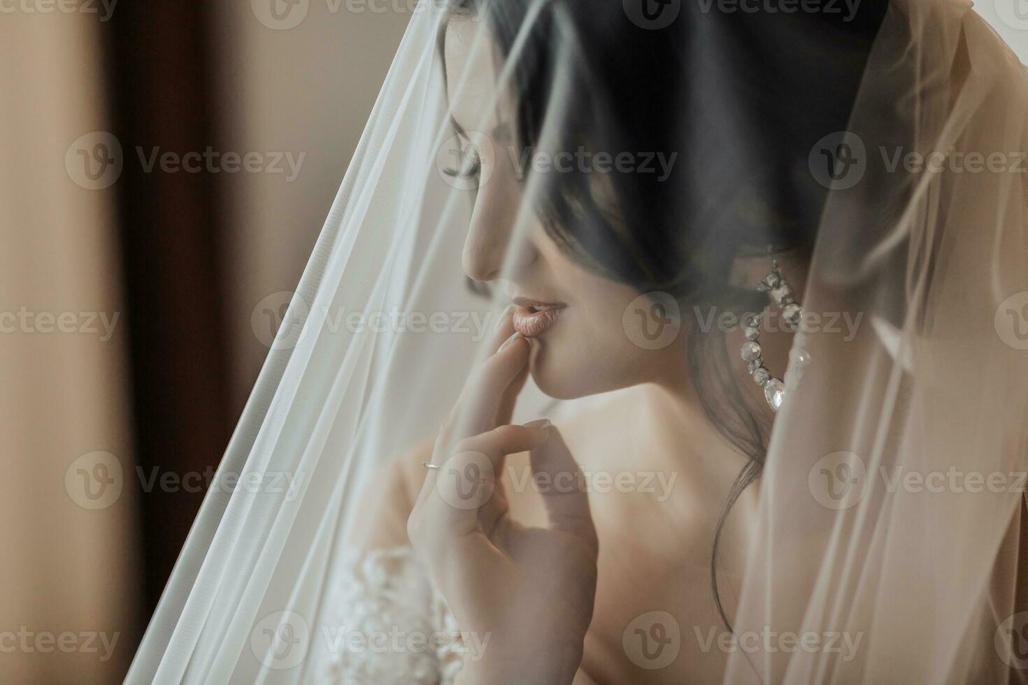 Portrait. A brunette bride in a white dress, wrapped in a veil, looks down and touches her face, poses. Gorgeous make-up and hair. Voluminous veil. Wedding photo. Beautiful bride photo