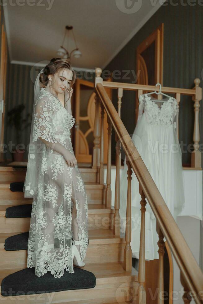 A delicate wedding dress hangs on a beautiful railing in the hallway of the house. The bride climbs the steps in a delicate transparent petticoat. Nice light. Wedding photo. A high quality photo