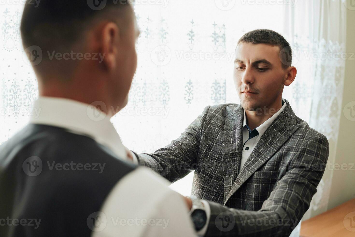 Photo of two men in classic suits. A handsome young man mends the shirt of another man standing in front of him. Business style. Stylish men. Clock