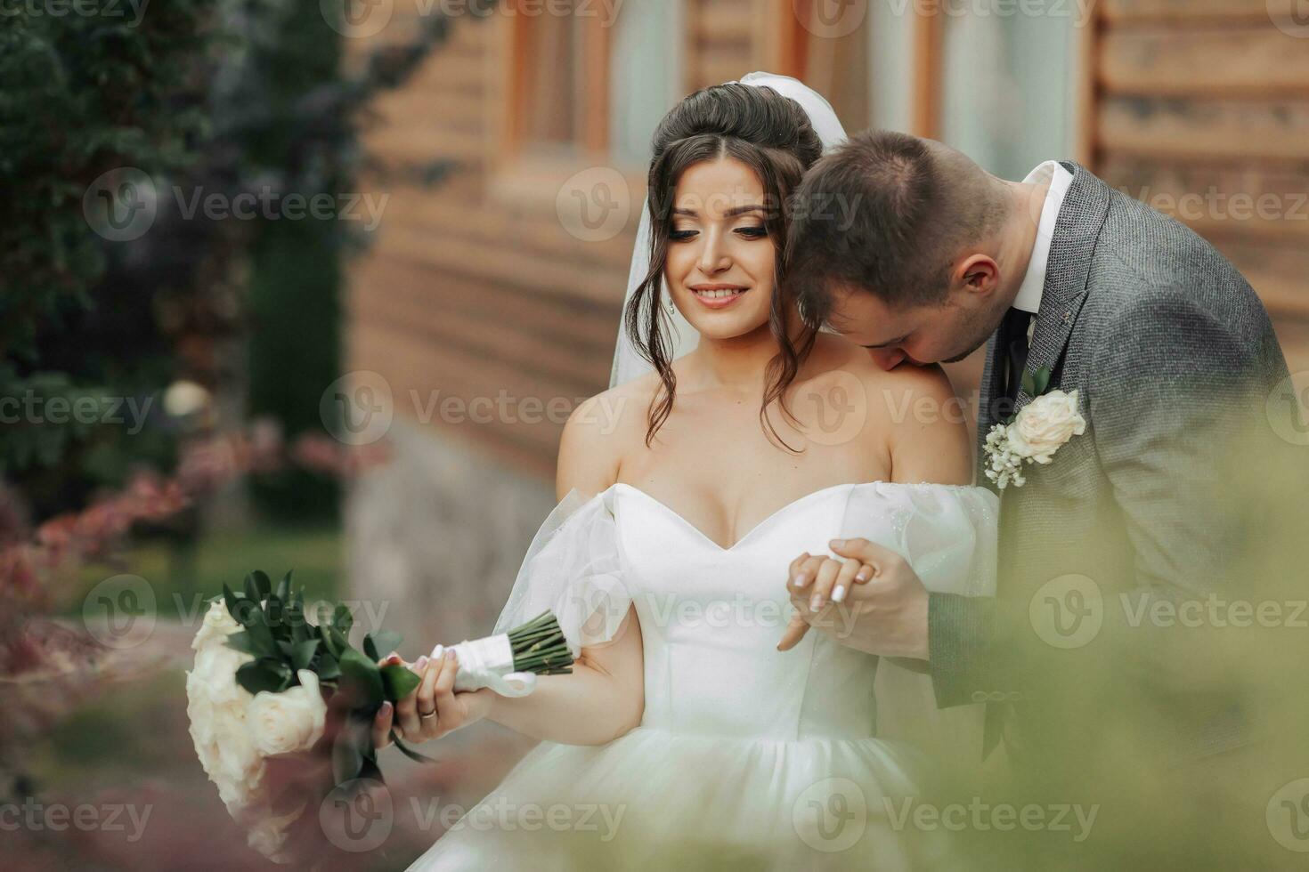 retrato de el novia y novio en naturaleza. un morena novia en un largo fuera del hombro blanco vestir y un velo y un elegante novio pose, el novio Besos su hombro. hermosa Rizado pelo y constituir. foto