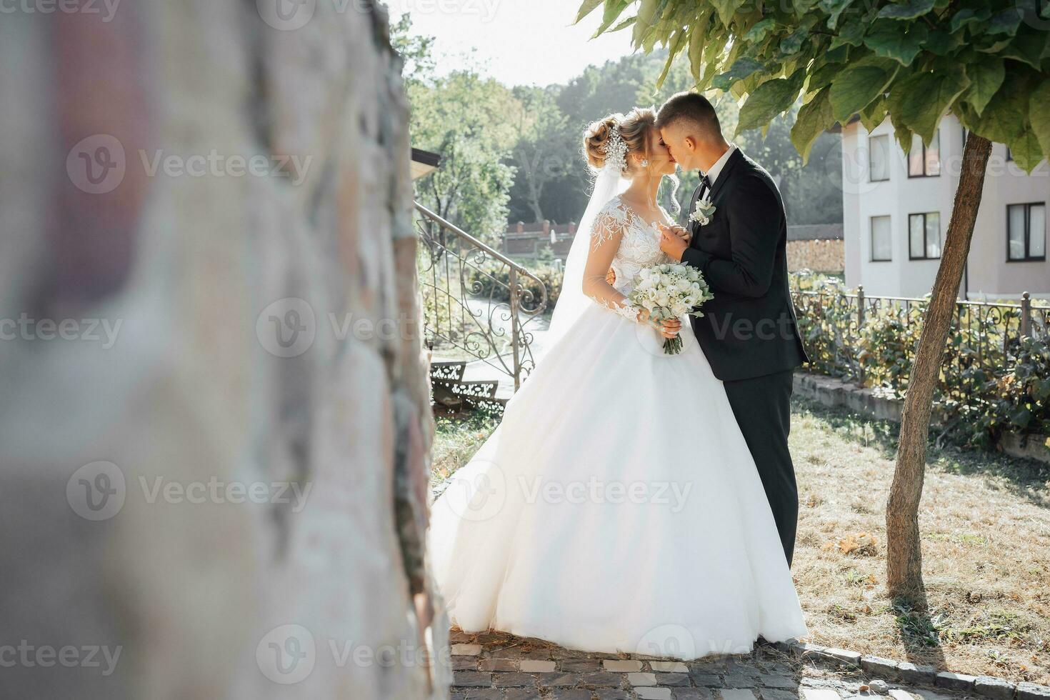 Wedding portrait. The groom in a black suit and the blonde bride stand by a stone wall under a tree, the groom kisses the bride's hand. Photo session in nature. Beautiful hair and makeup