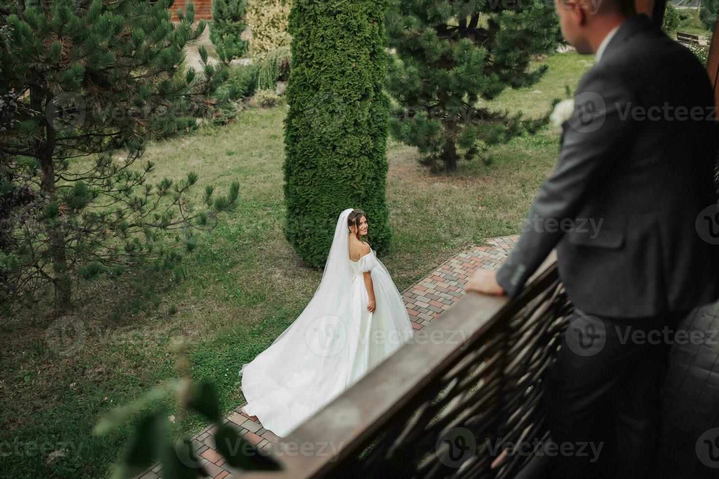 Wedding photo. The bride in a voluminous white dress and a long veil walks in the garden along a stone path, the groom stands on the balcony in the foreground, his shoulders turned to her. photo