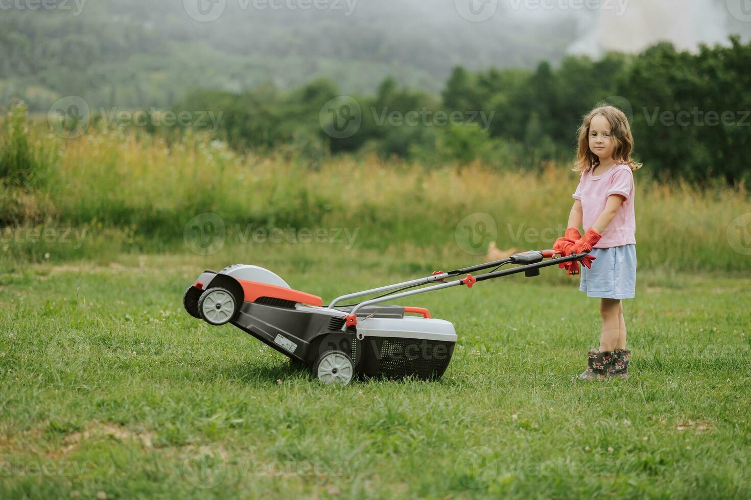 A child in boots in the form of a game mows grass with a lawnmower in the yard against the background of mountains and fog, the concept of garden tools photo