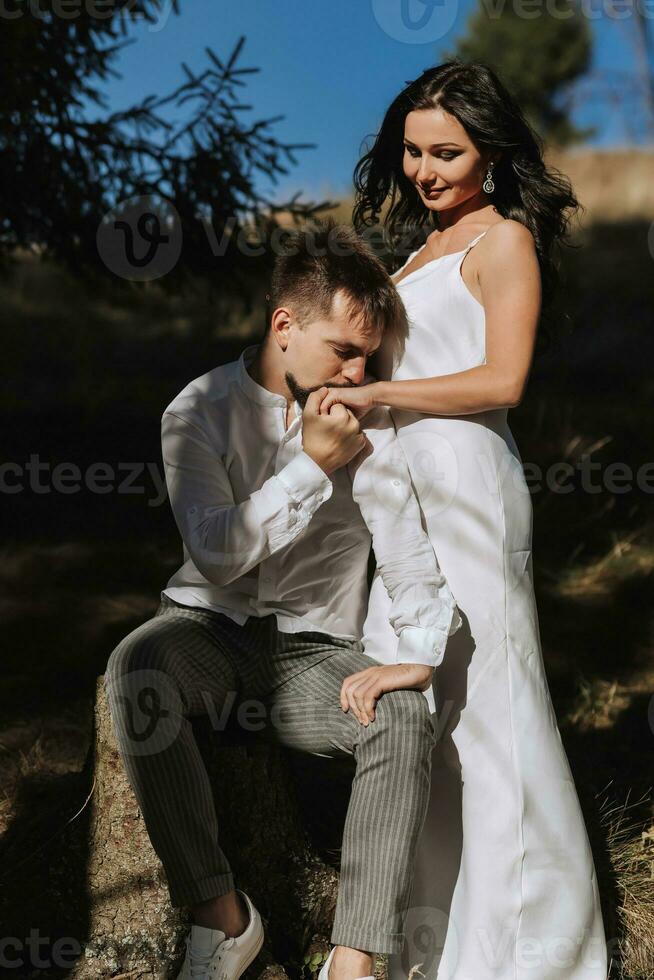 Stylish model couple in the mountains in summer. A young girl in a white silk dress stands next to her husband against the background of a forest and mountain peaks. photo