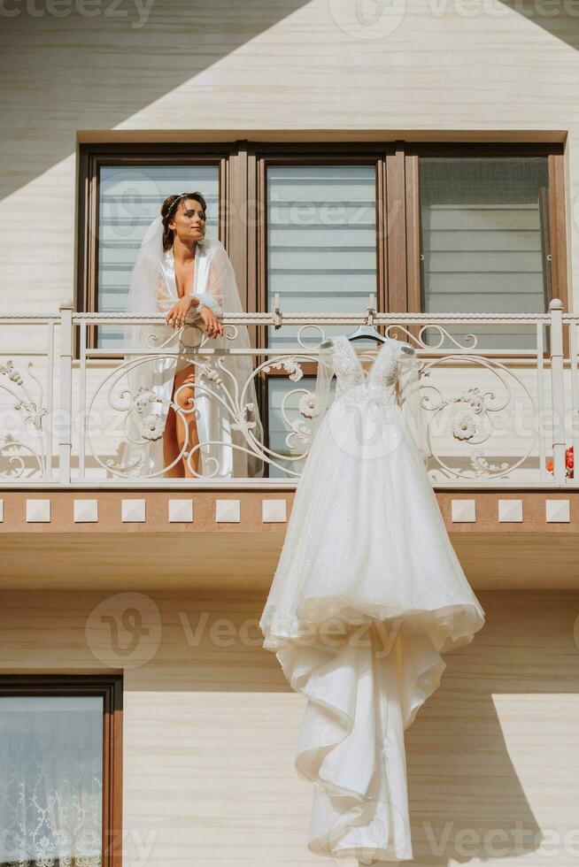 white wedding dress hanging on balcony railing, wide shot of house in front and girl on balcony. Wedding details, modern wedding dress with long train, long sleeves and open back photo