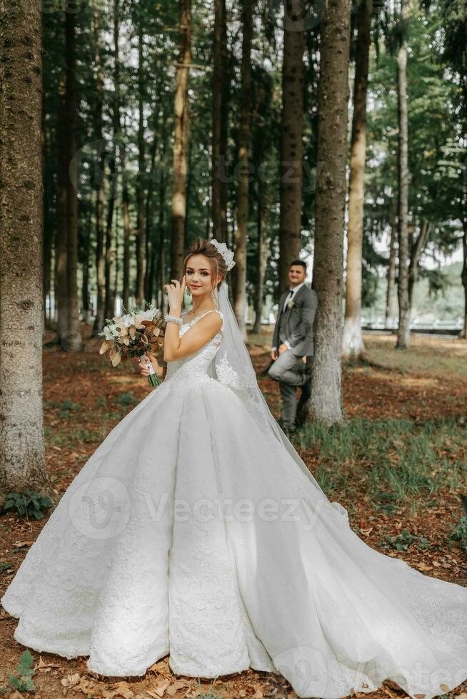 A beautiful young woman in a wedding dress between tall trees in the forest with a royal hairstyle and a chic tiara with a bouquet of flowers in her hands, a wedding in golden color photo