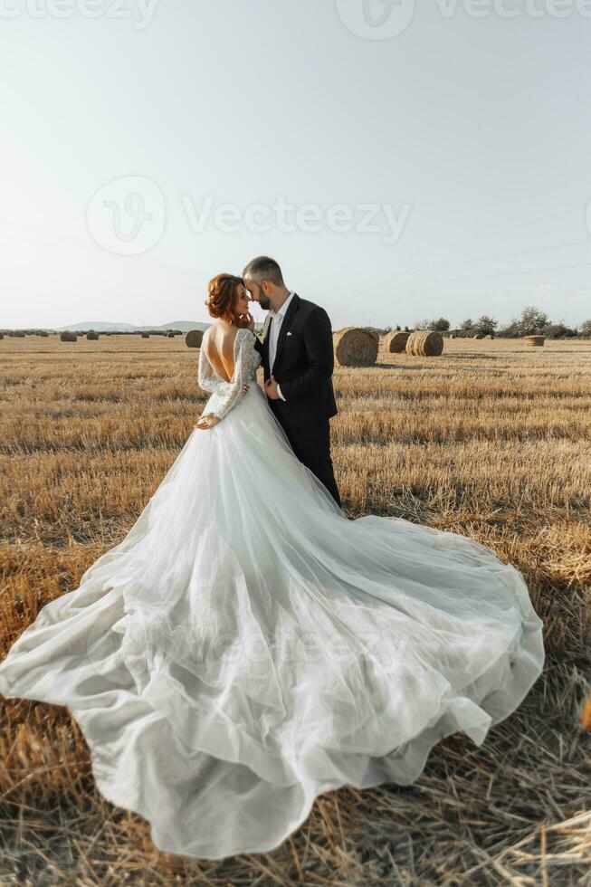 The bride and groom are standing in the field, and behind them are large sheaves of hay. The bride stands with her shoulders turned to the camera. Long elegant dress. Stylish groom. Summer photo