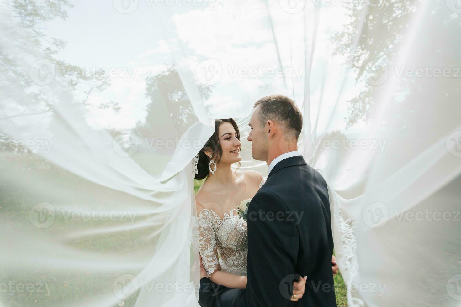 A curly-haired brunette bride in a white dress and a veiled groom embrace and kiss. Portrait of the bride and groom. Beautiful makeup and hair. Wedding in nature photo