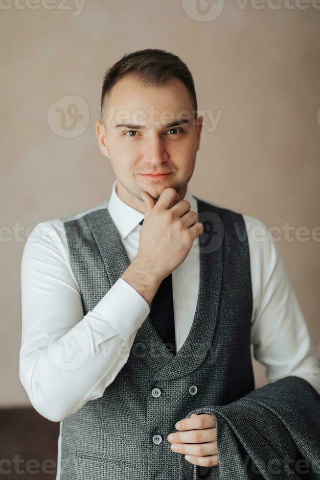 A man in a white shirt and a gray vest is posing by the window and looking at the camera, holding his jacket. Front view. A stylish watch. Men's style. Fashion. Business photo