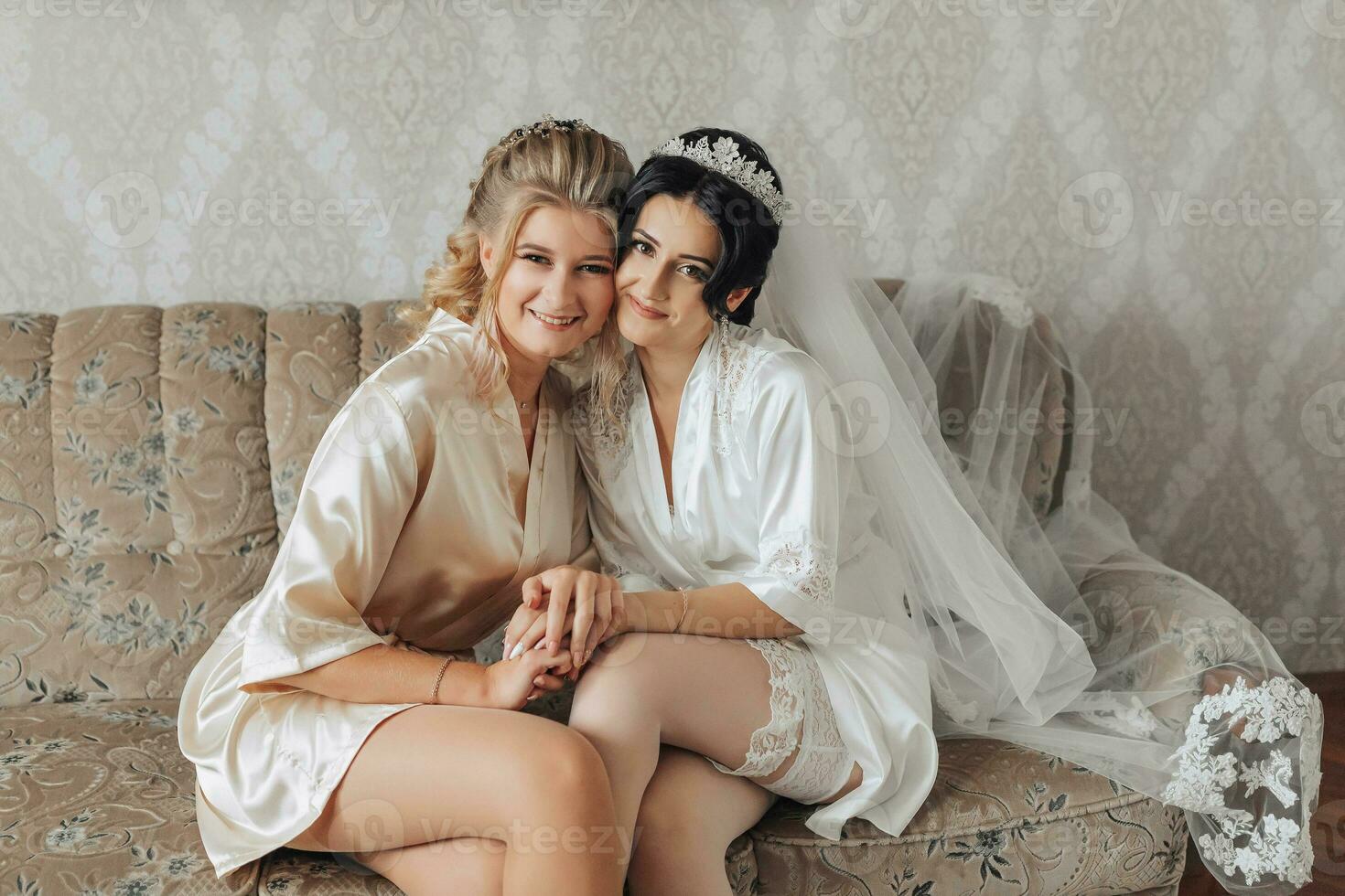 A brunette bride and her friend are sitting on a sofa in the bride's room, dressed in robes, smiling and posing. Wedding portrait wedding photo in a light key. Cropped photo. Morning of the bride.