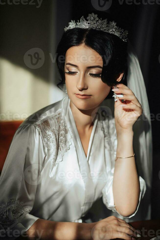 Portrait of a brunette bride sitting in a petticoat and touching her face. The silver crown is decorated with steel flowers. Gorgeous make-up and hair. Voluminous veil. Wedding photo. Beautiful bride photo