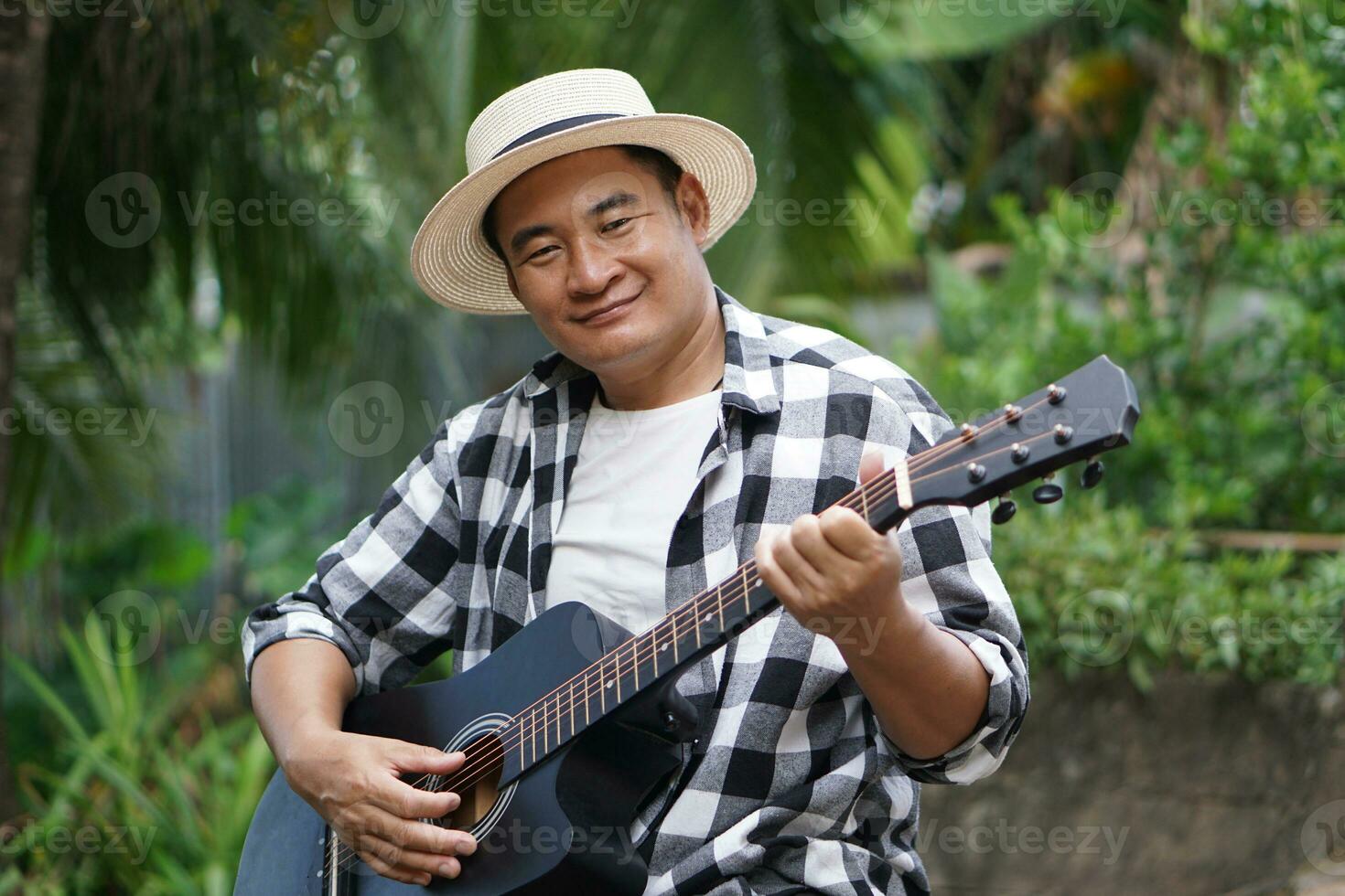 Handsome Asian man is playing acoustic guitar in the park, outdoor nature background. Concept, love music, hobby, recreation activity. Relax time. Music can develop mental health. photo