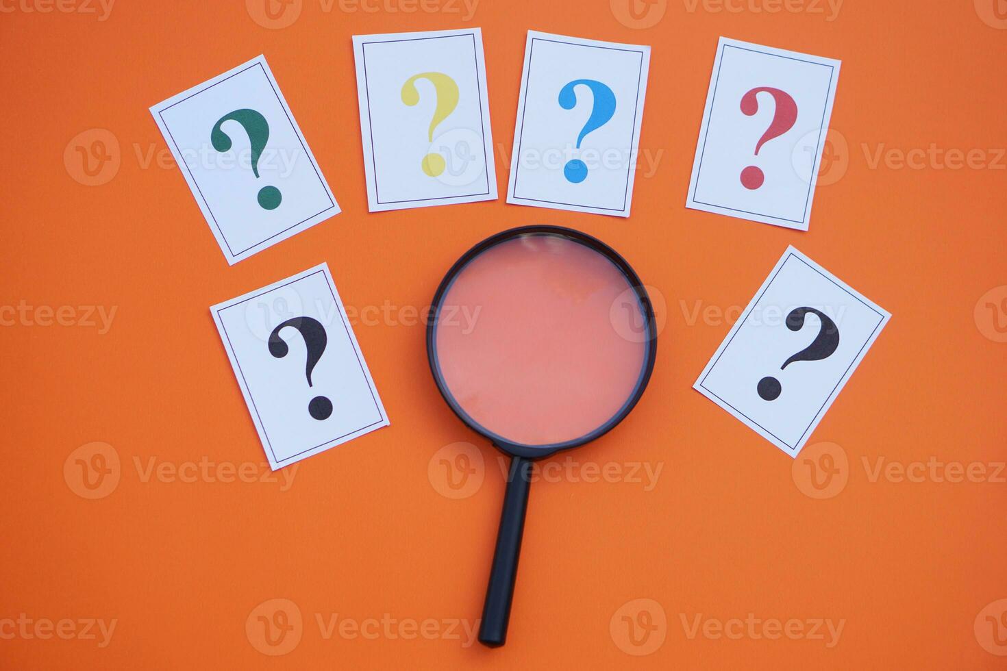 Paper cards of colorful question marks and magnifying glass on orange background. Concept. Teaching aid. Education materials for doing activity or playing investigation games about finding answers. photo