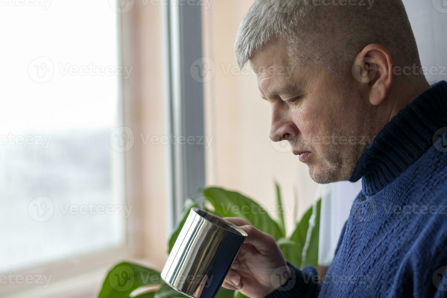 Portrait of the mid aged man with grey hair, wearing warm, dark blue sweater, drinking coffee. People photo