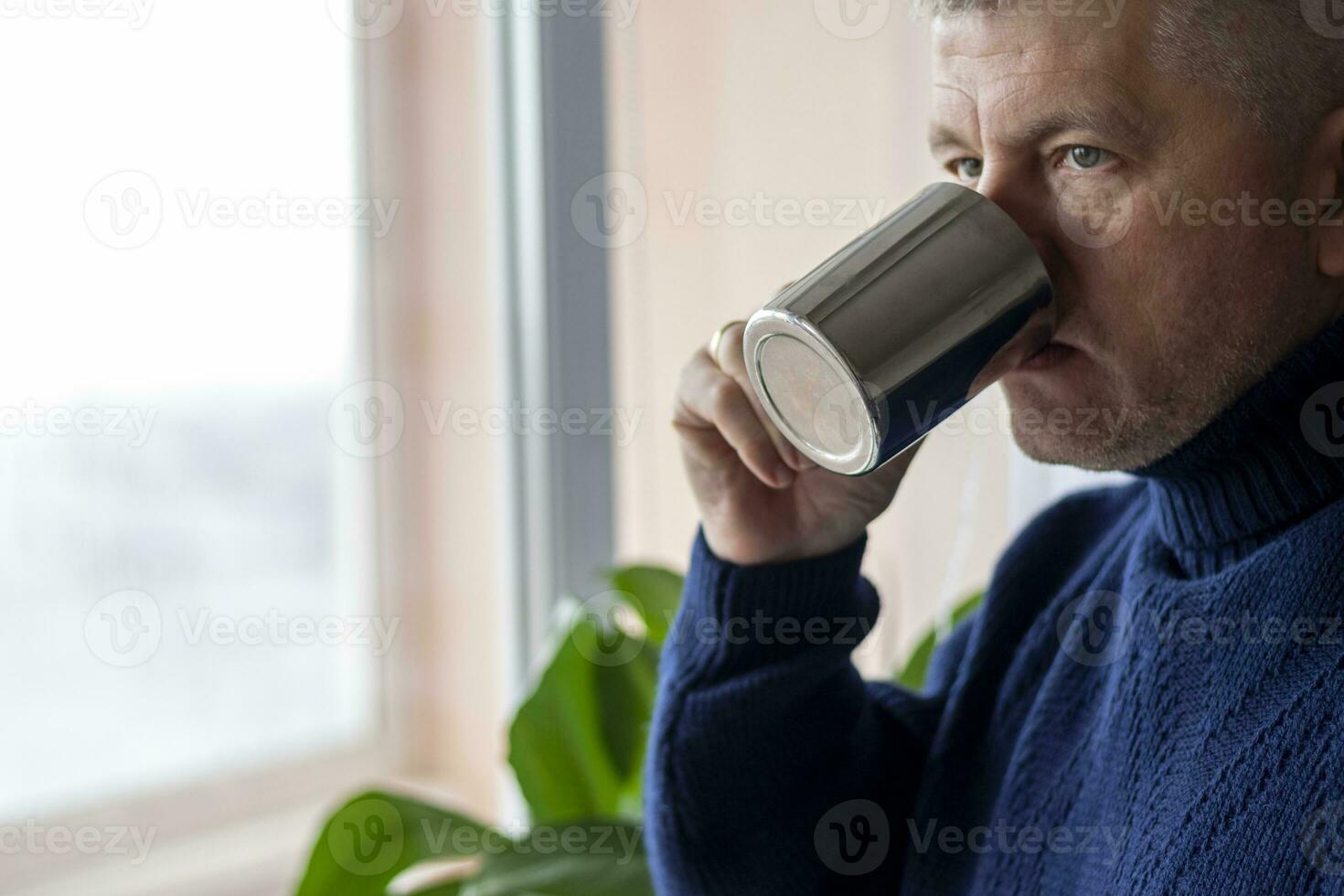 Portrait of the mid aged man with grey hair, wearing warm, dark blue sweater, drinking coffee. People photo