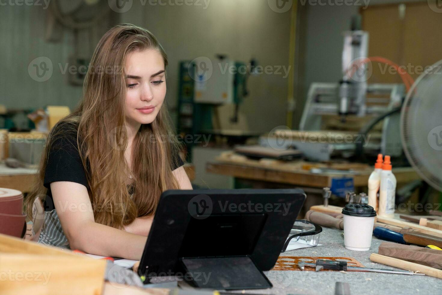 A young woman is training to be a carpenter in the workshop. She works with a laptop computer in a wood workshop. female carpenter contact customers by smartphone. SME orders, Start-up and small photo