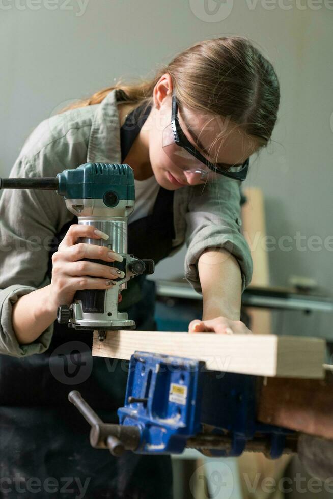 hembra carpintero vistiendo protector la seguridad lentes y utilizando eléctrico trabajo en un madera. artista o mueble diseñador trabajando en un producto idea en un taller. foto