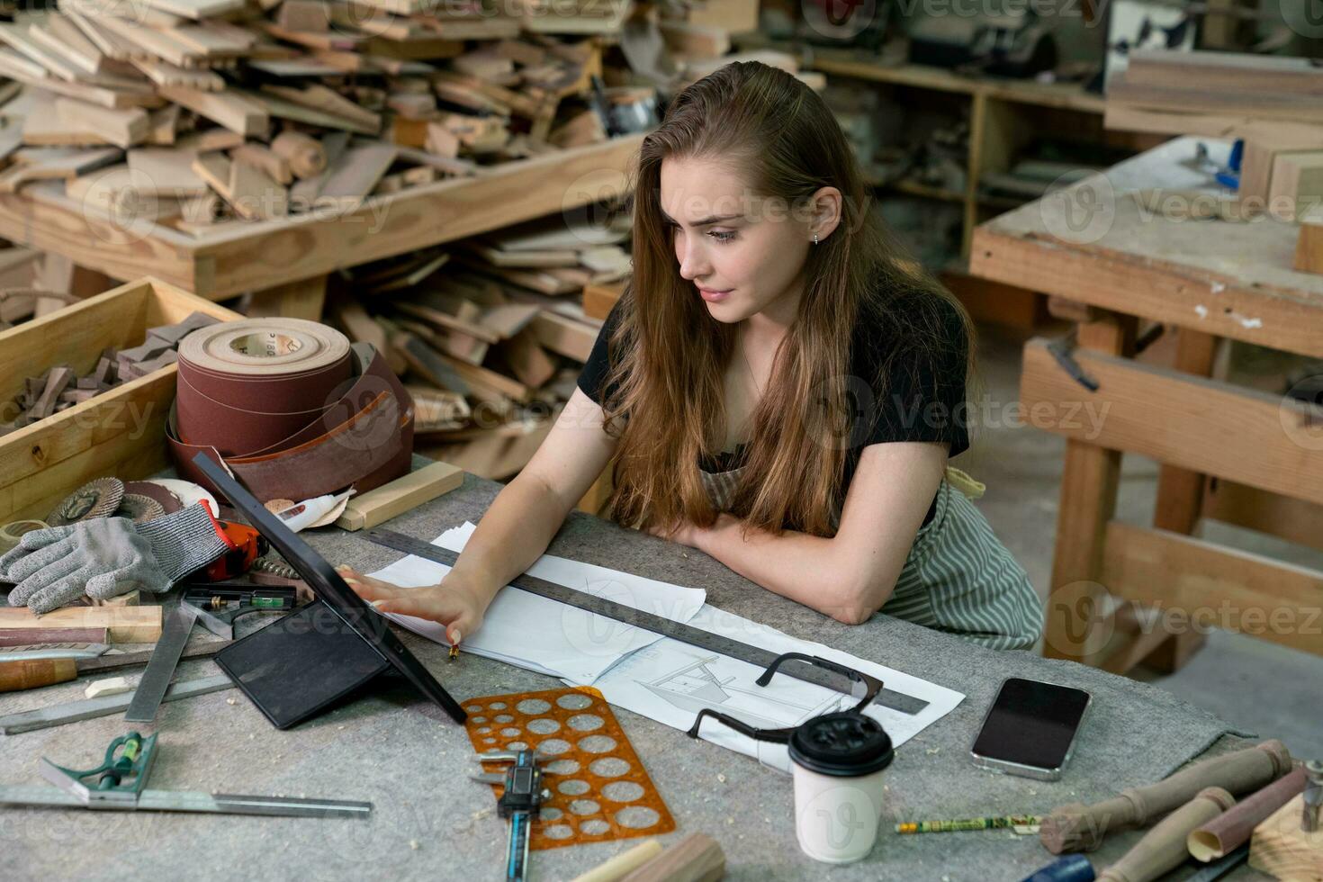 A young woman is training to be a carpenter in the workshop. She works with a laptop computer in a wood workshop. female carpenter contact customers by smartphone. SME orders, Start-up and small photo