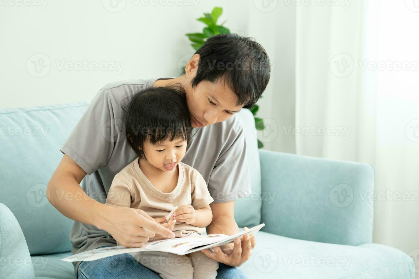 Happy Asian father relax and read book with baby time together at home. parent sit on sofa with daughter and reading a story. learn development, childcare, laughing, education, storytelling, practice. photo