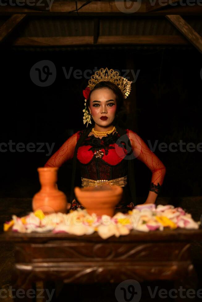 An Asian woman in a red dancer's dress is sitting at a ritual offering table full of flowers photo