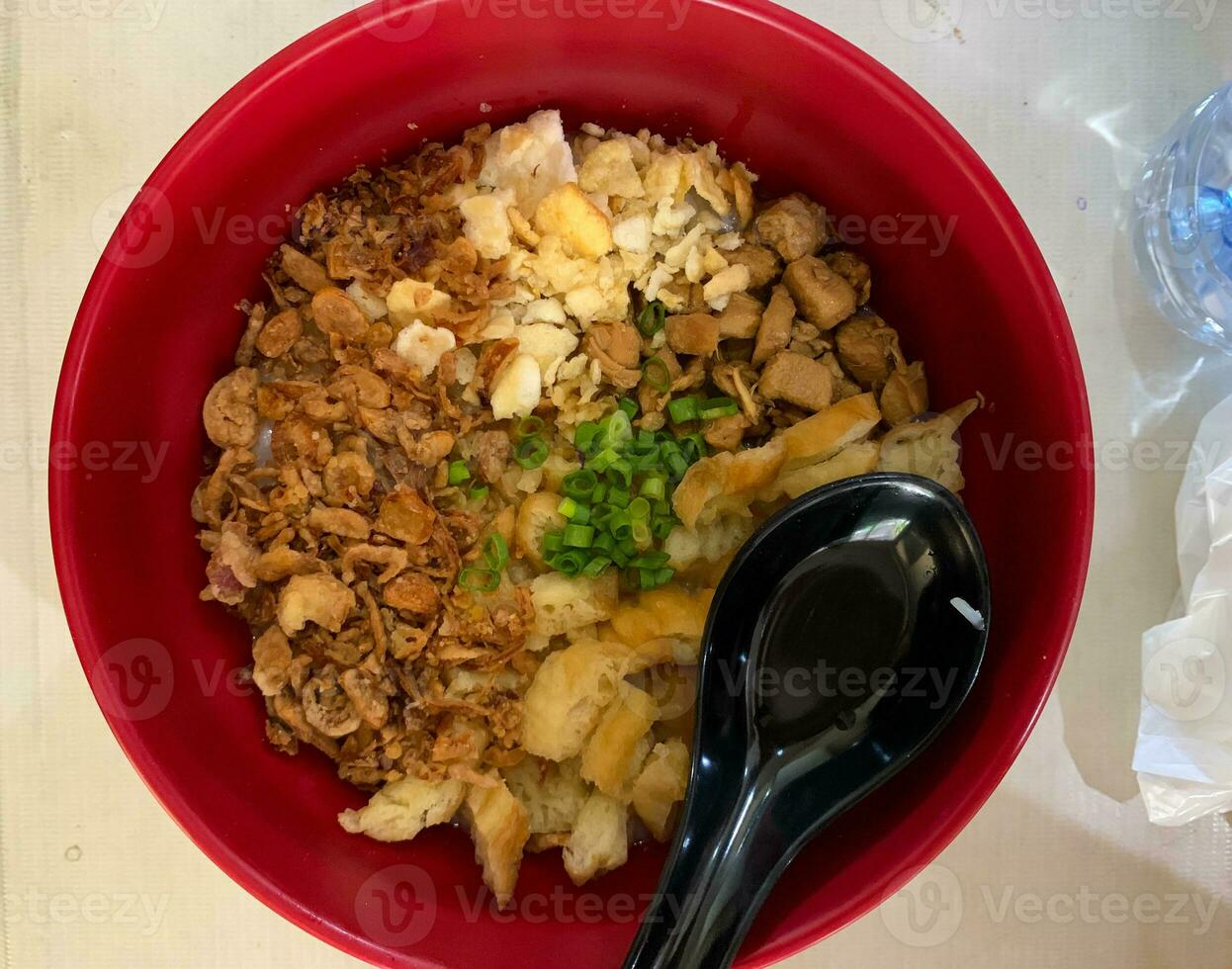 closeup bubur ayam or chicken rice porridge in red bowl with black spoon and topping, top view photo